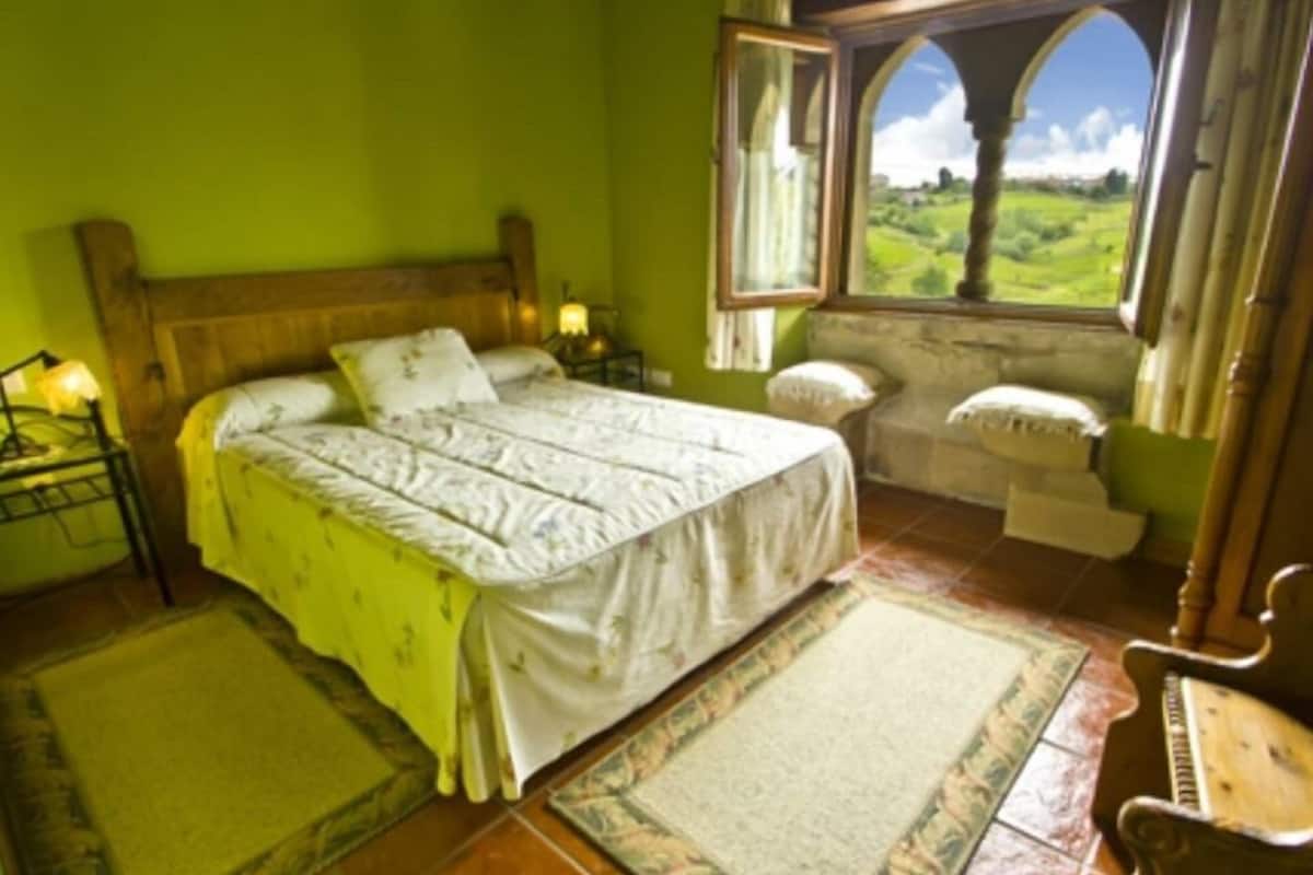 Un dormitorio con una cama grande, una mesita de noche, una silla, un ventanal con vistas al campo y un cabecero de madera.