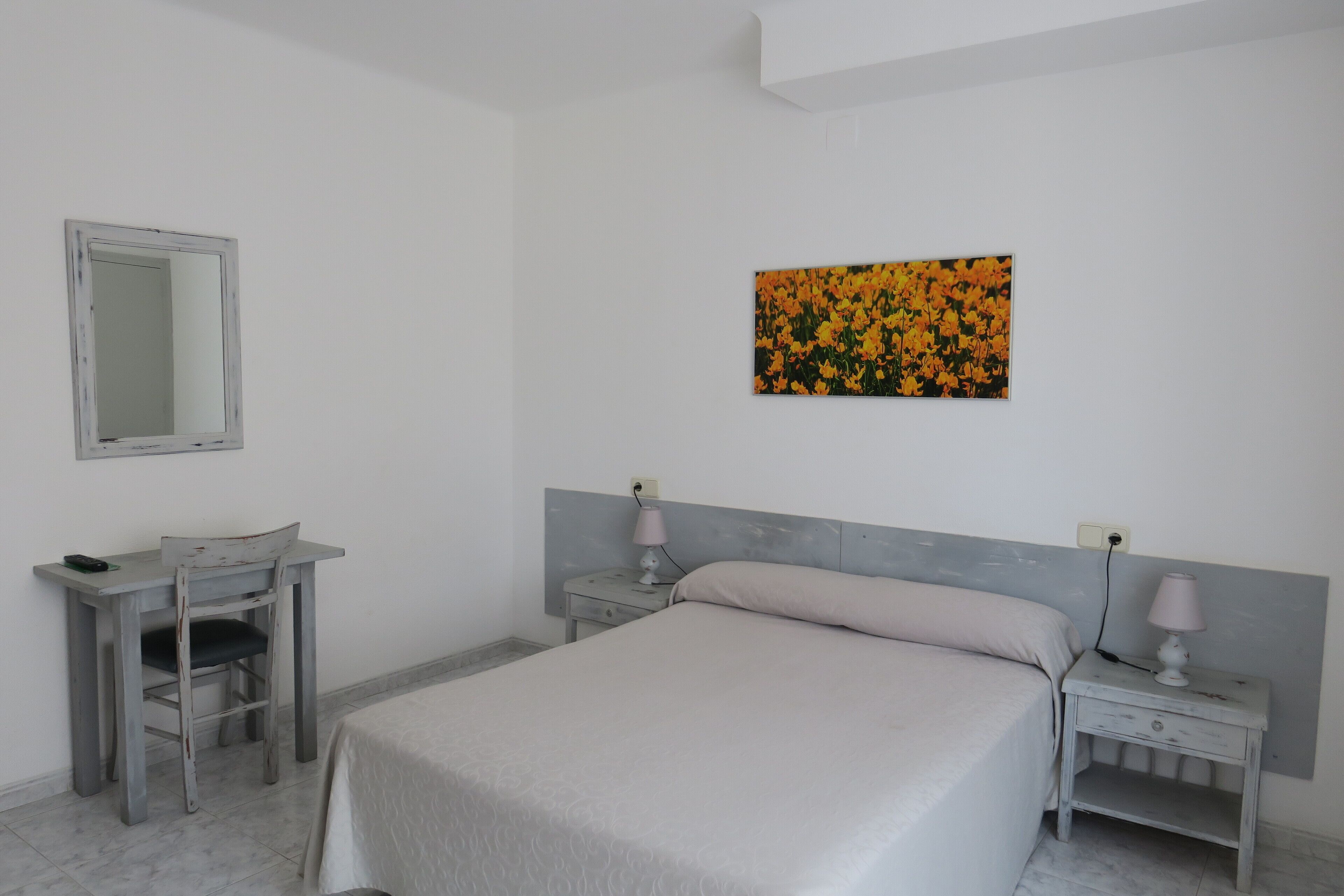 A bedroom with a bed, a desk, a chair, a nightstand, a lamp, a mirror, and a painting on the wall.