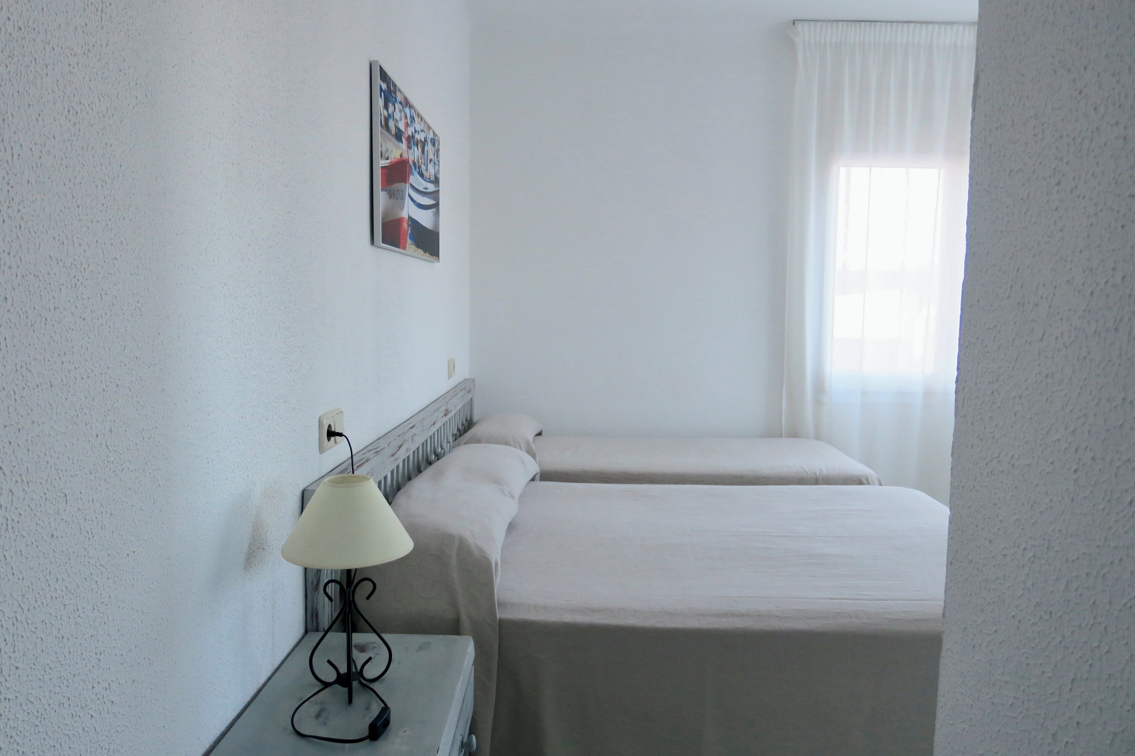 Ein Hotelzimmer mit zwei Betten, einem Nachttisch mit Lampe, einem Bild an der Wand und einem Fenster mit Vorhängen.