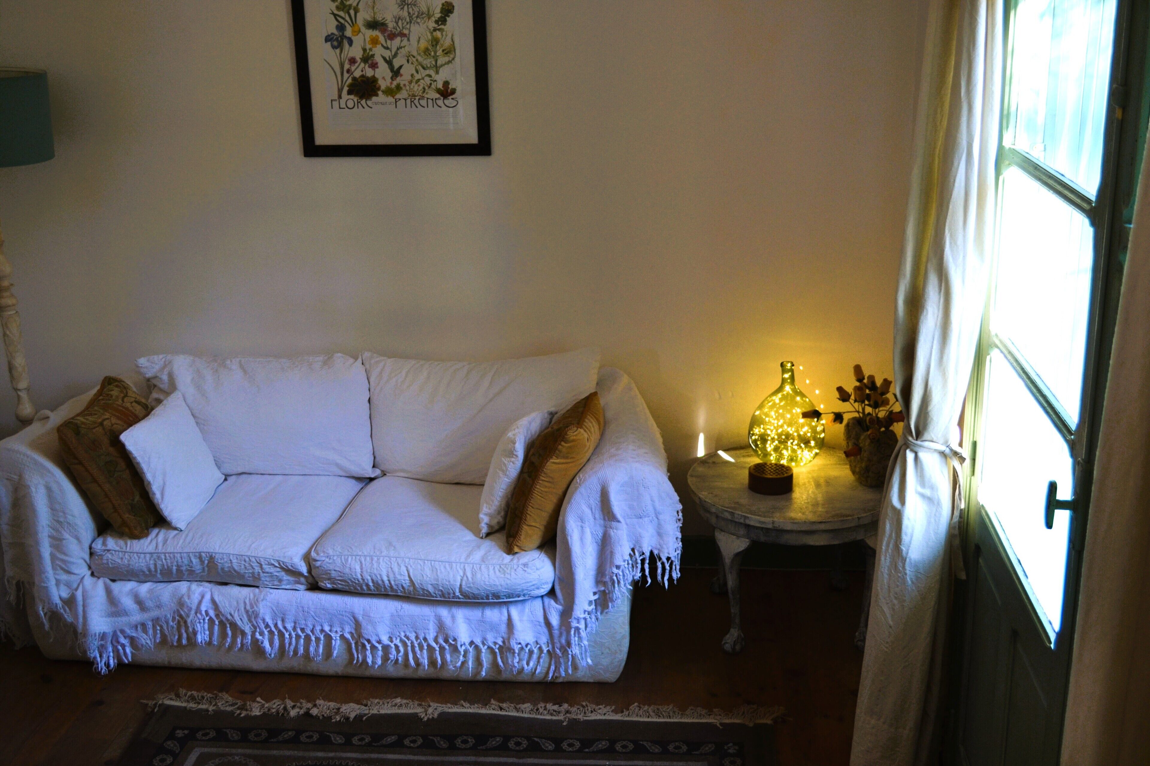 Un salon chaleureux avec un canapé blanc, une petite table d’appoint avec une lampe et une fenêtre avec des rideaux.
