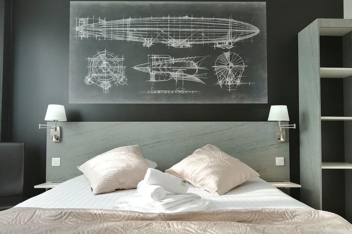A hotel room with a bed, pillows, a folded towel, and a wall-mounted artwork of a zeppelin and other aircraft.