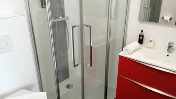 Un baño moderno con ducha de vidrio, lavamanos blanco con detalles rojos y un espejo sobre el tocador.