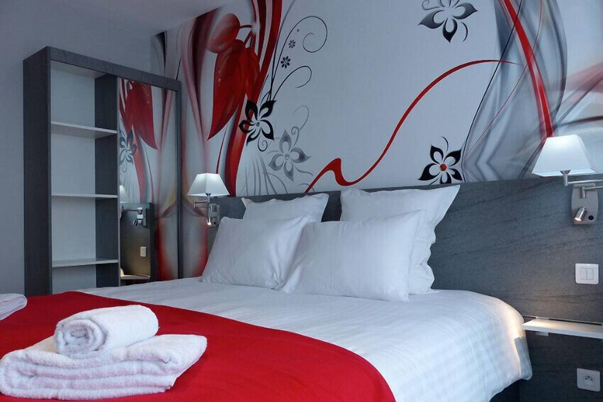 A modern hotel room with a large bed, white and red bedding, a grey headboard, and a wall with floral and abstract designs.