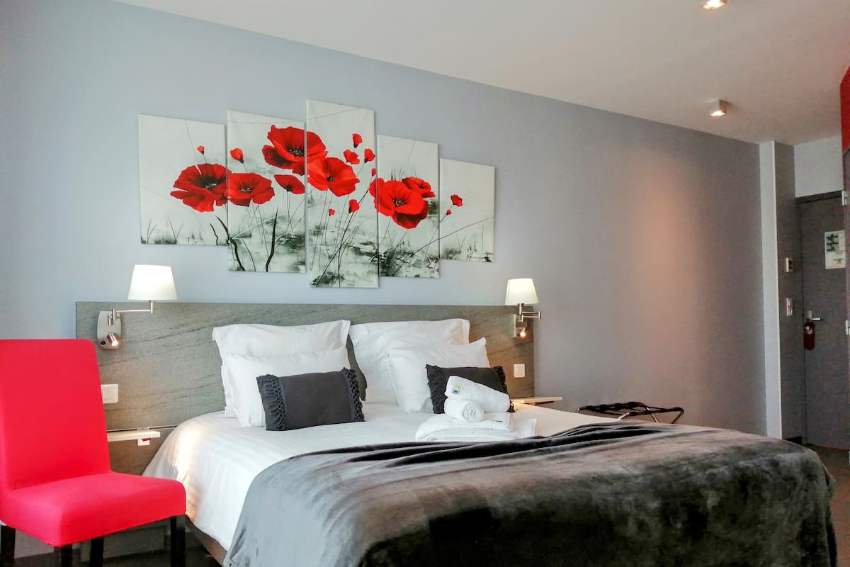 A hotel room with a bed, a red chair, a large painting of red flowers, and two bedside lamps.