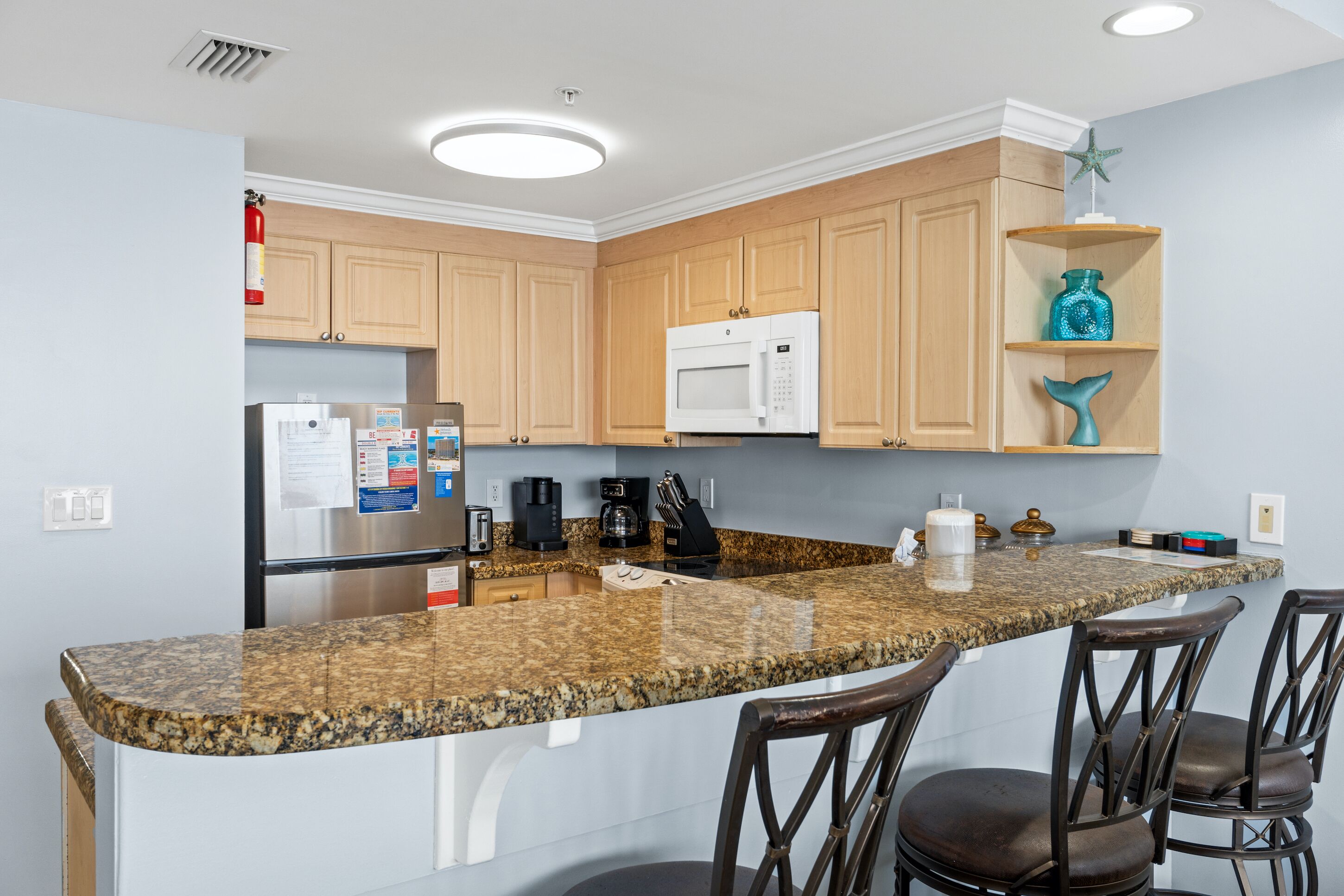 A kitchen with granite countertops, wooden cabinets, a stainless steel refrigerator, a microwave, a coffee maker, and a starfish decoration on a shelf.