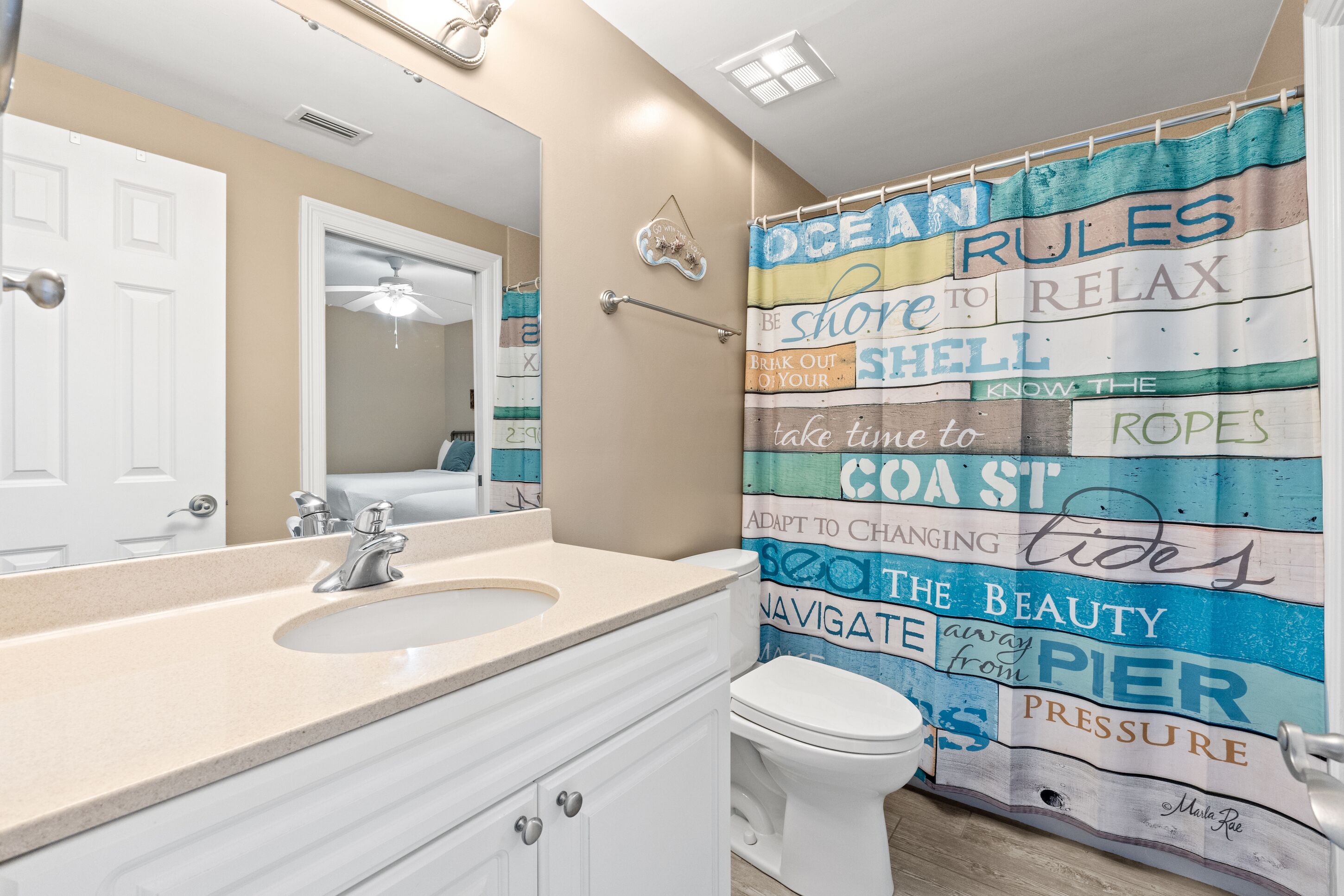 A bathroom with a beige countertop, a white sink, a white toilet, and a shower curtain with nautical-themed text.