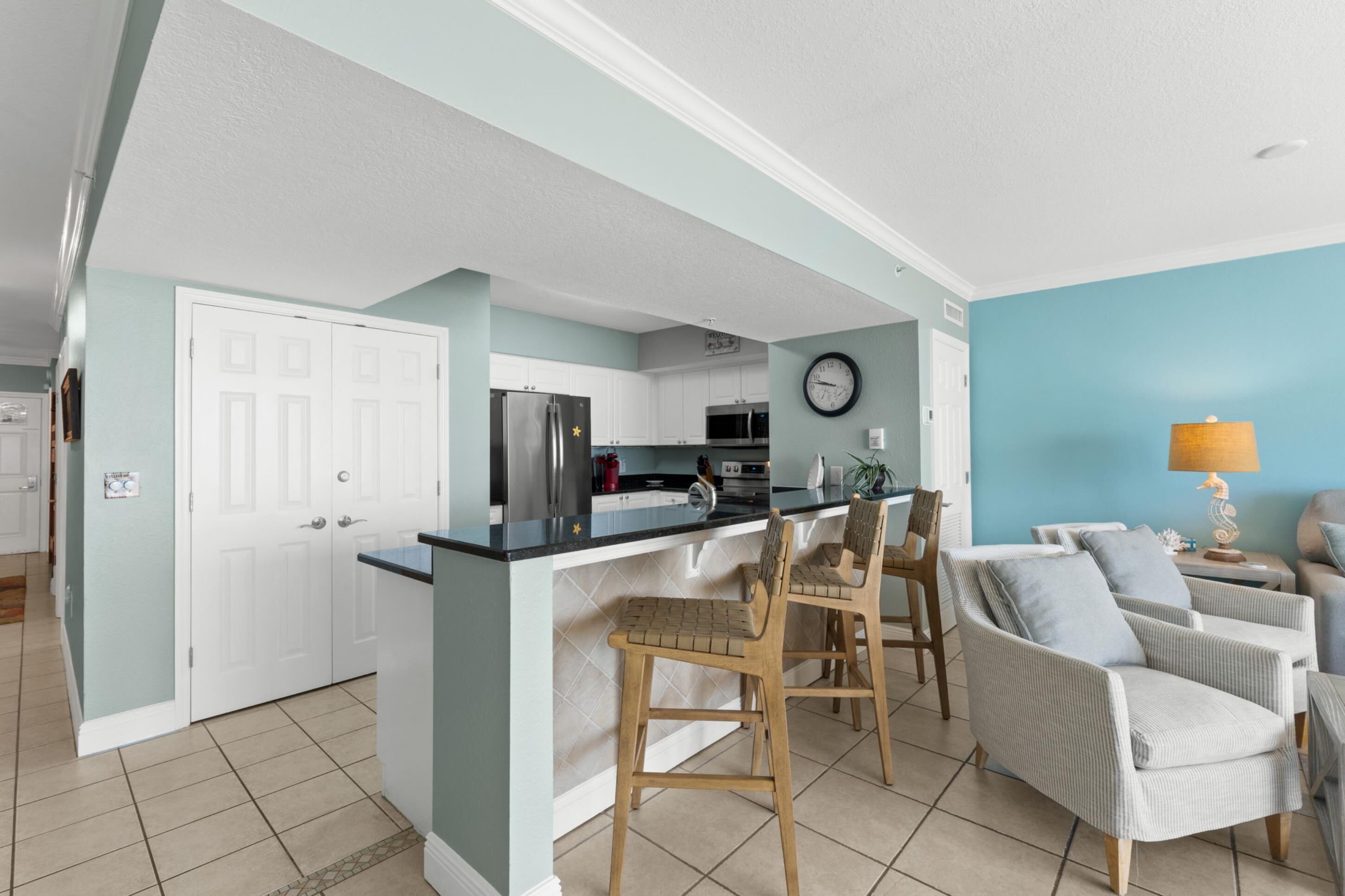 A modern kitchen with white cabinets, a black countertop, and a stainless steel refrigerator. There are two wooden chairs and a white armchair in the dining area.