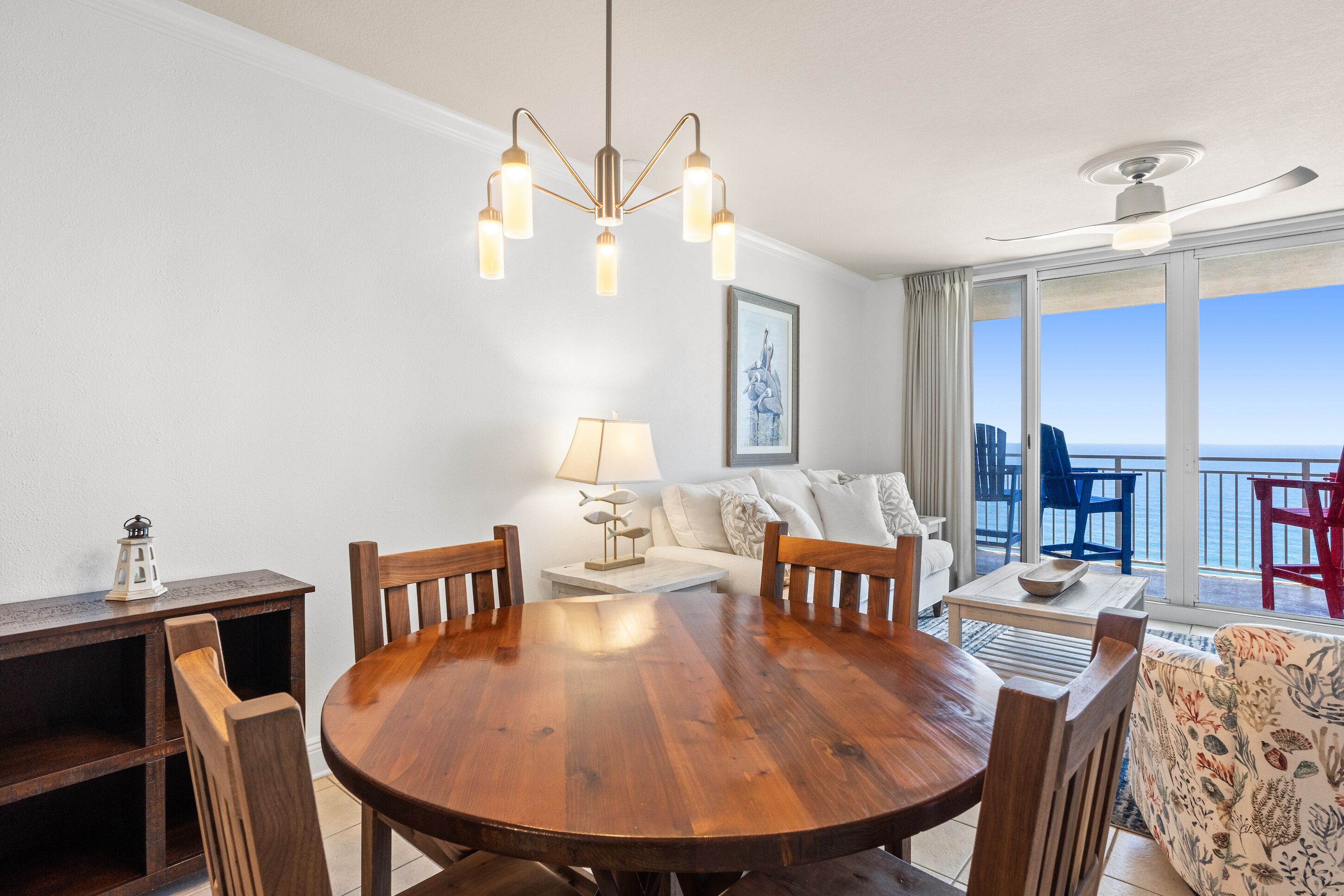 A dining area with a wooden table and chairs, a chandelier, a sofa, and a view of the ocean through large windows.