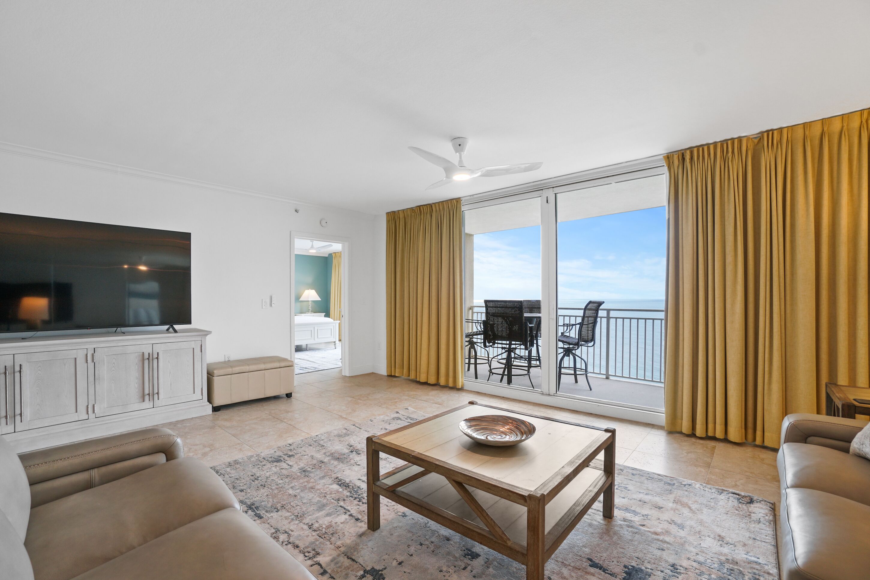 A spacious living room with a flat-screen TV, a wooden coffee table, and a sofa. A balcony with a view of the ocean is visible through large sliding doors.