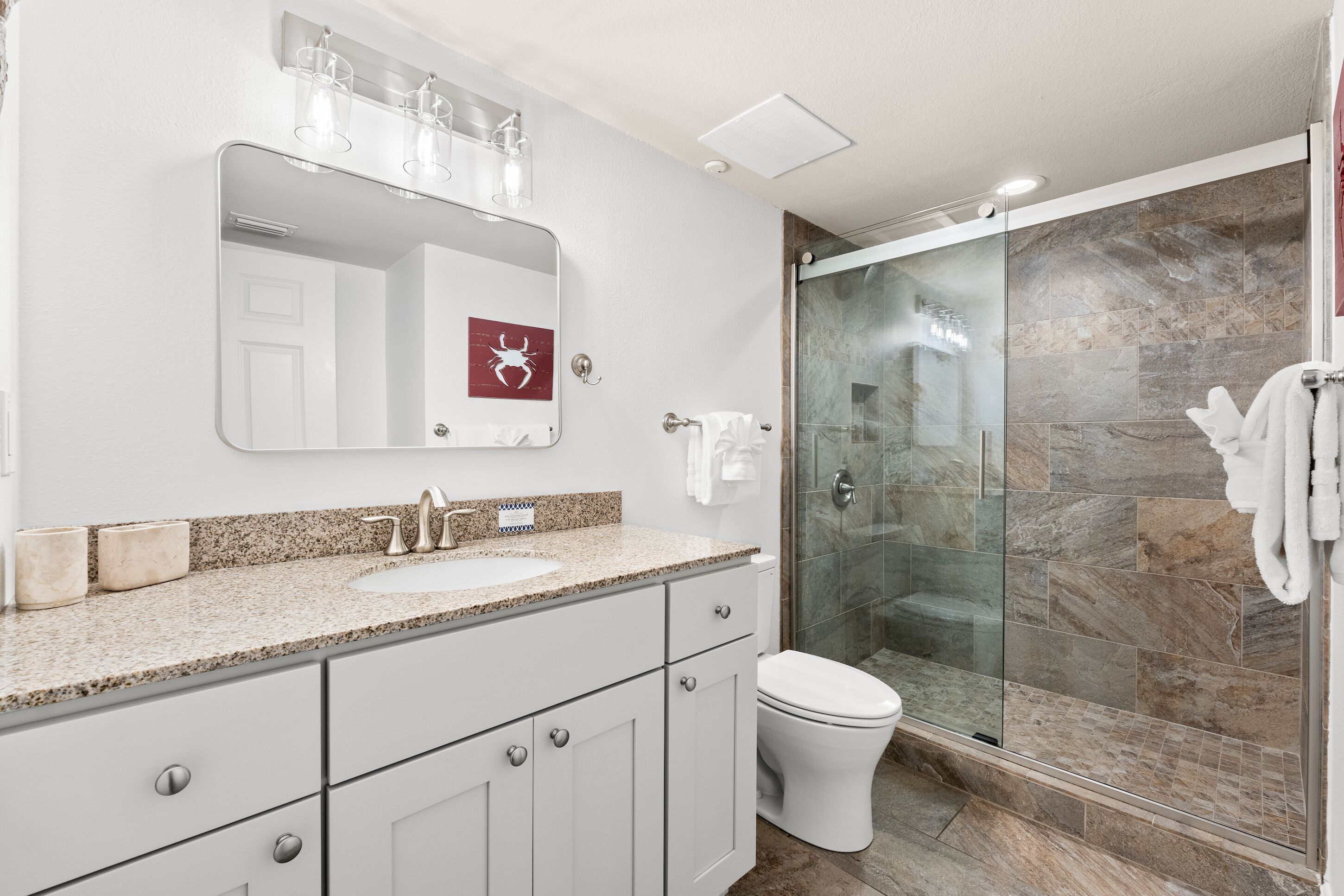 A modern bathroom with a granite countertop, a white toilet, a glass-enclosed shower, and a mirror with a red symbol.