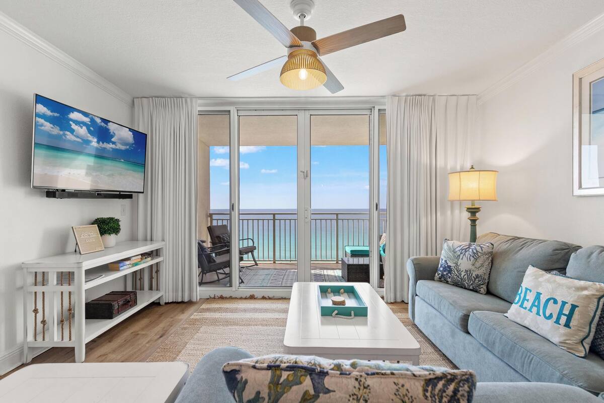 Un salon équipé d’une télévision, d’un ventilateur de plafond, d’un canapé agrémenté d’un coussin orné du mot « PLAGE », d’une table basse et d’une porte-fenêtre coulissante donnant sur un balcon avec vue sur l’océan.