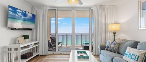 Ein Wohnzimmer mit Fernseher, Deckenventilator, einer Couch mit einem 'BEACH'-Kissen, einem Couchtisch und einer Glasschiebetür, die auf einen Balkon mit Meerblick führt.