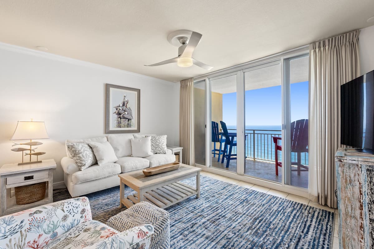 Un salon moderne avec un canapé, une table basse et un téléviseur. Il y a un balcon avec vue sur l’océan.