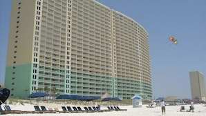 Een groot hotel met meerdere verdiepingen en balkons, nabij een strand met ligbedden en parasols.