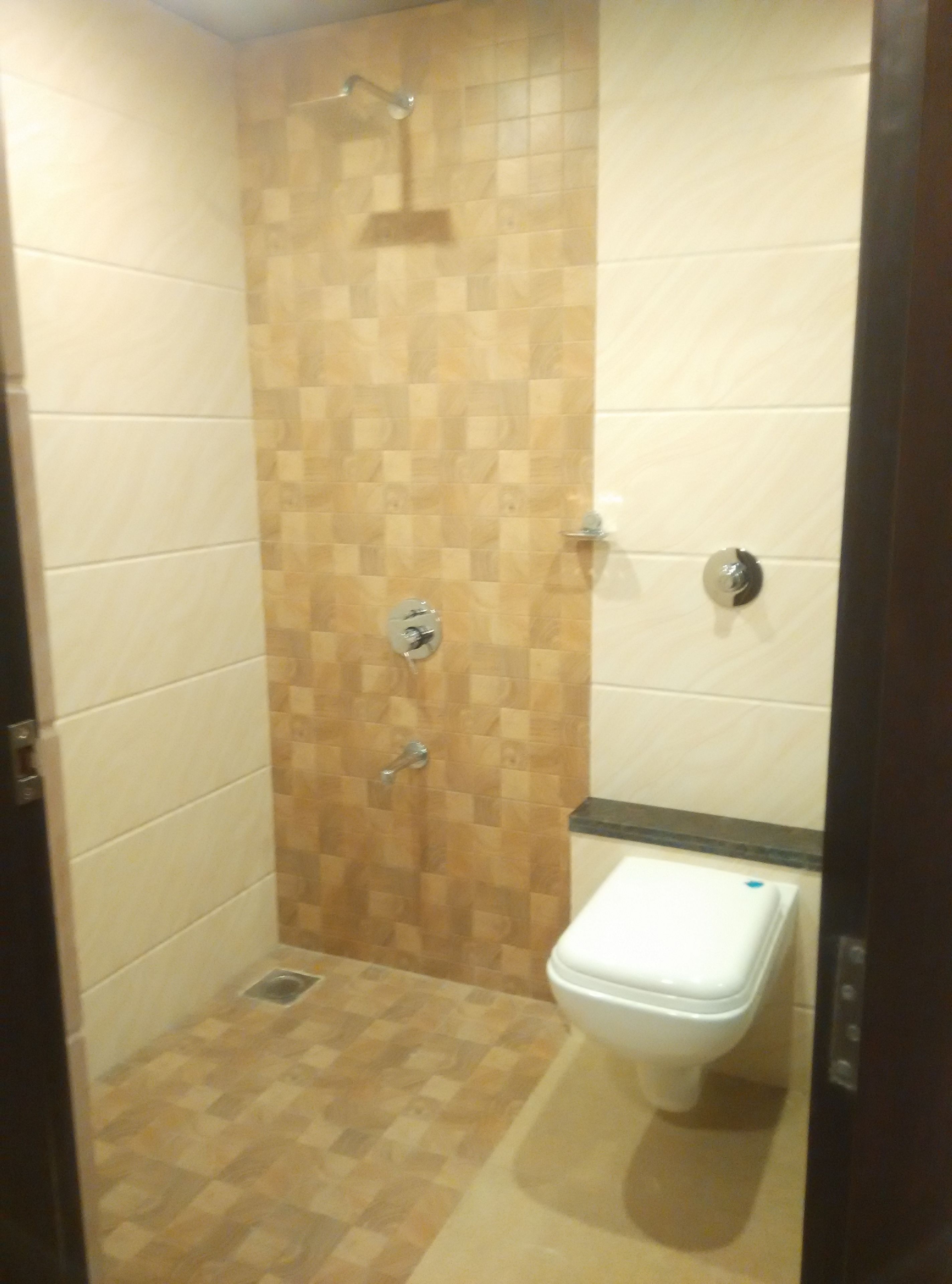 Un pequeño box de ducha cerrado con pared y suelo de baldosas a cuadros, un cabezal de ducha manual y un inodoro con asiento blanco.