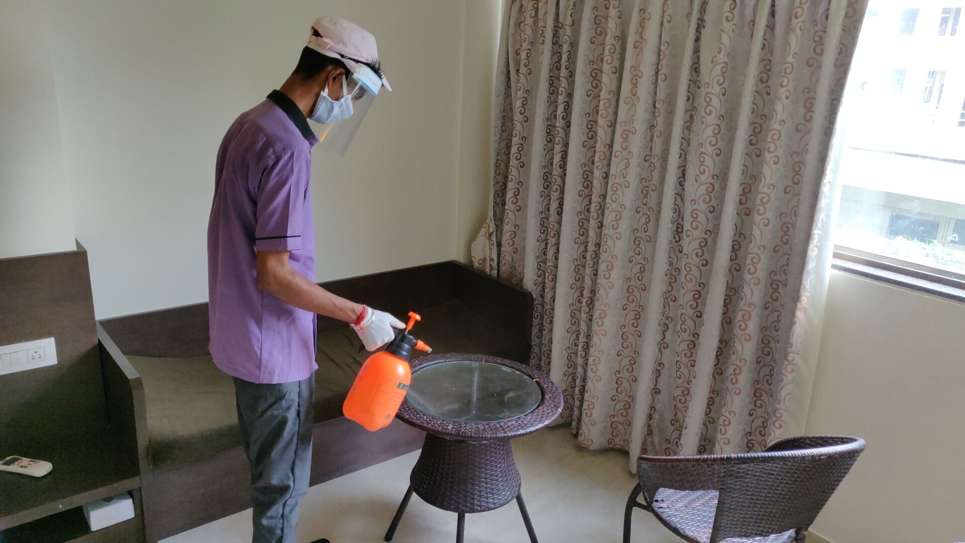 Uma pessoa vestindo camisa e boné roxos limpando uma mesa de vidro com um borrifador.