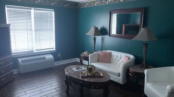 A room with teal walls, a white sofa, a white armchair, a round wooden table with a tray, two lamps, and a large window with blinds.