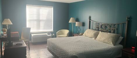 A bedroom with a large bed, two bedside tables, a chair, a desk, and a window with blinds.
