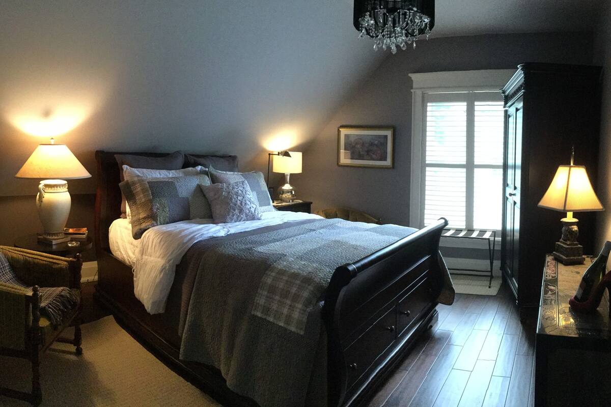 Un dormitorio ordenado con una cama, dos mesitas de noche con lámparas, una silla y una ventana con persianas.
