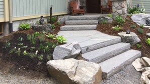 Una casa con entrada de piedra, un porche con sillas y un jardín con escalones que llevan a la puerta.