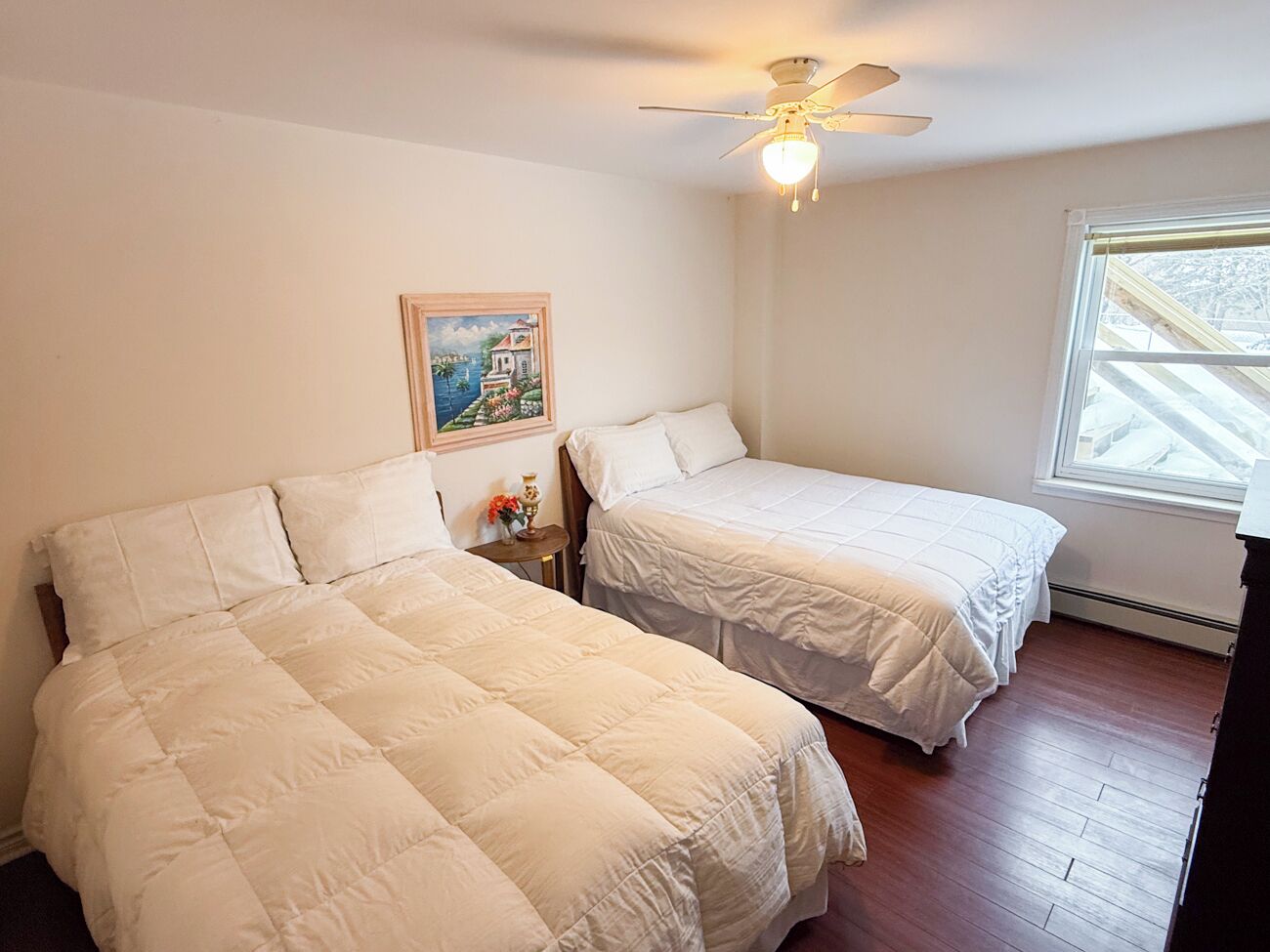 Une chambre avec deux lits, un ventilateur de plafond, une fenêtre avec des stores et un tableau au mur.