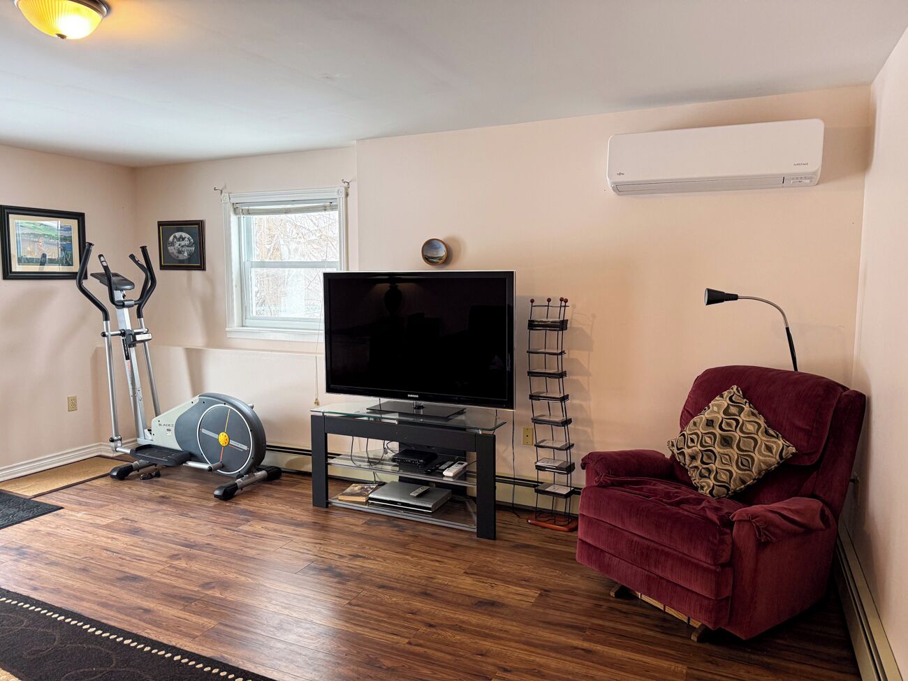 Een woonkamer met een loopband, een flatscreen televisie op een glazen standaard, een rode fauteuil en een raam met jaloezieën.