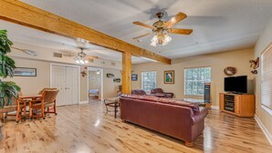 A spacious living room with hardwood flooring, a leather sofa, a dining table with chairs, a ceiling fan, and a television on a wooden cabinet. - The Chassahowitzka Hotel (Homosassa)