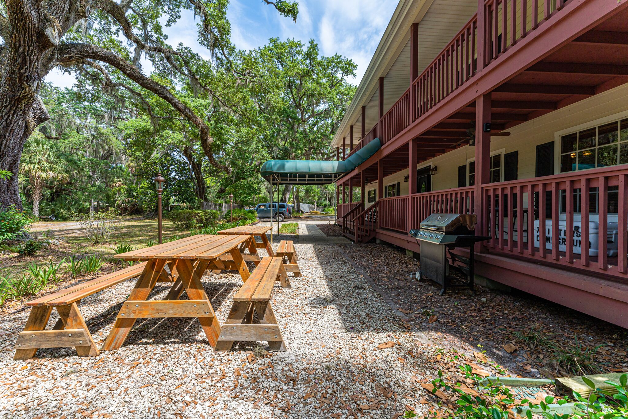 A covered picnic area with wooden benches and a grill, surrounded by trees and a building with a red balcony.
