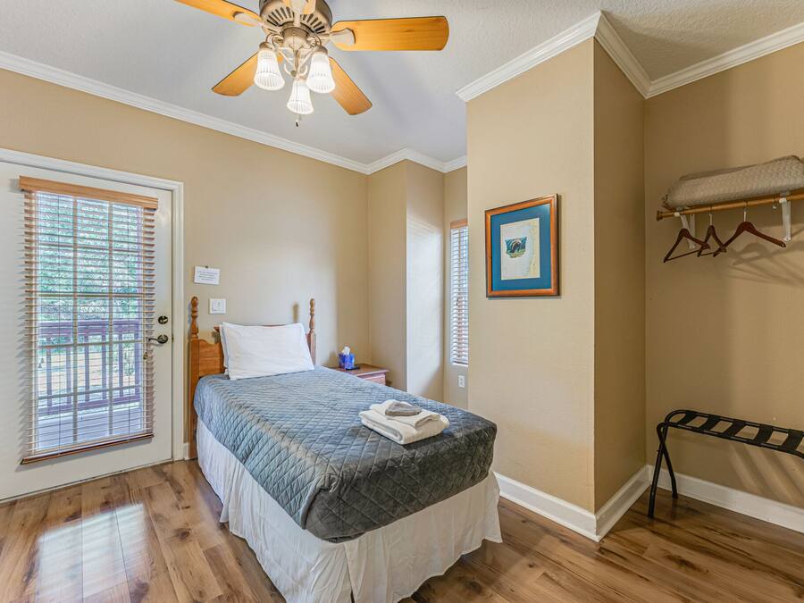 A bedroom with a bed, a ceiling fan, a door with blinds, a bedside table, and a wall-mounted shelf with a blanket.