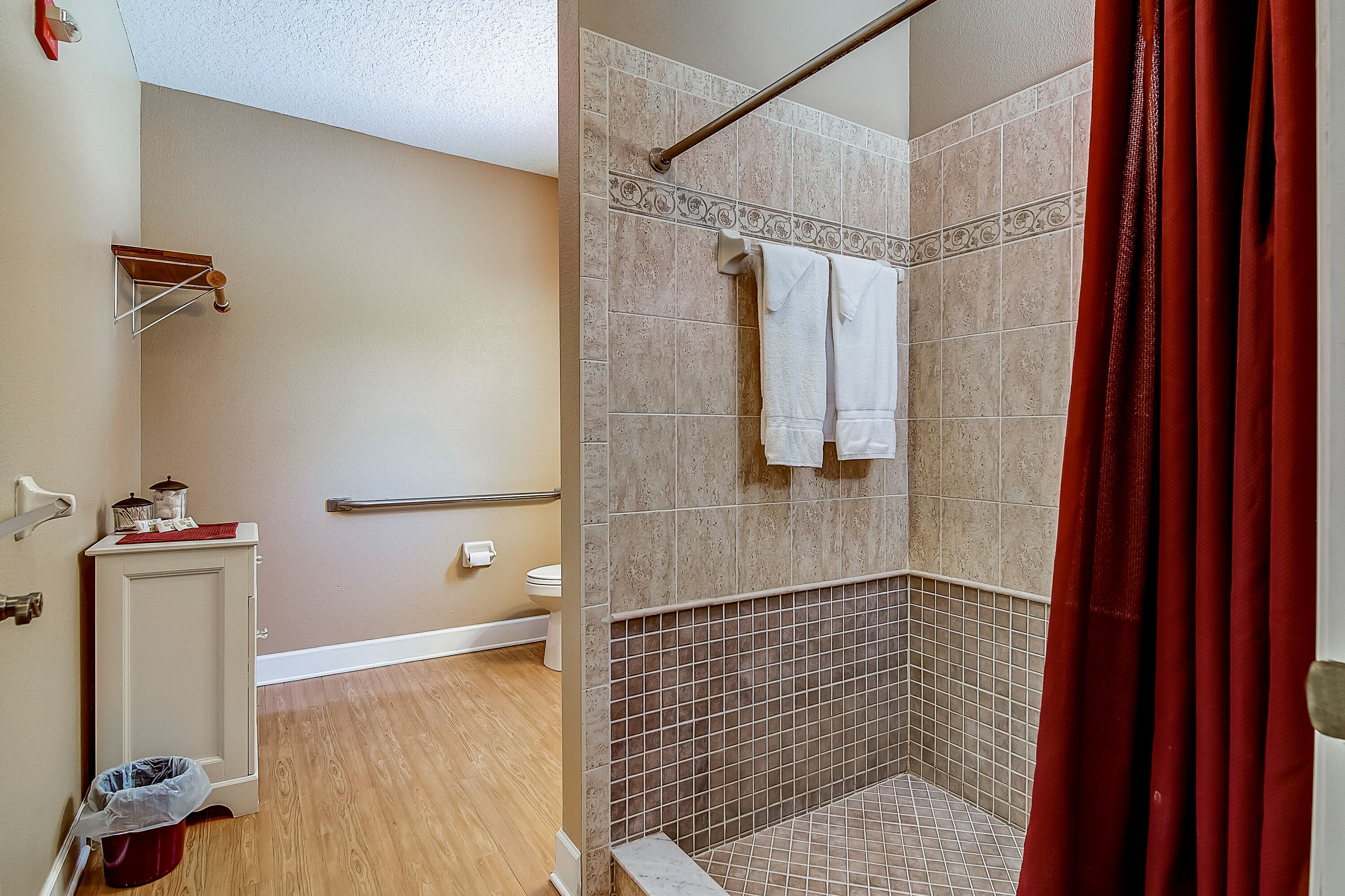 A bathroom with a shower, tiled walls, a wooden floor, a cabinet, and a red curtain.