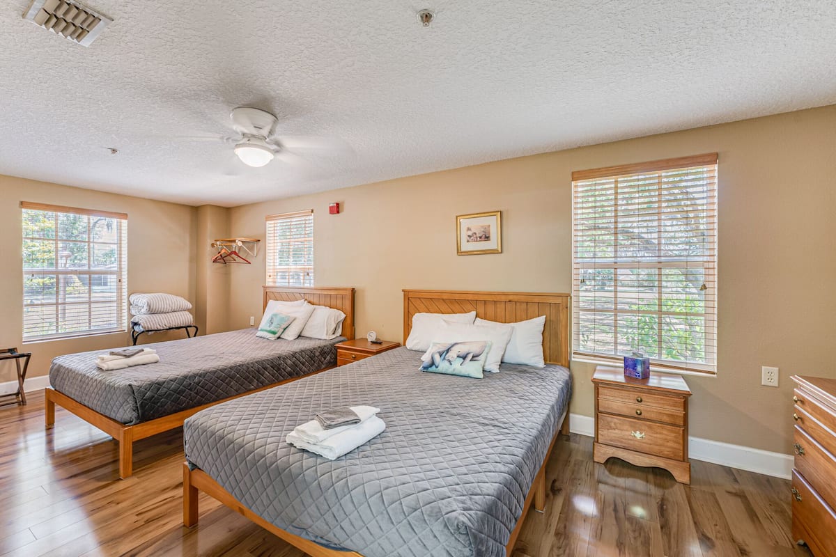 A room with two beds, wooden flooring, a ceiling fan, and large windows with blinds.