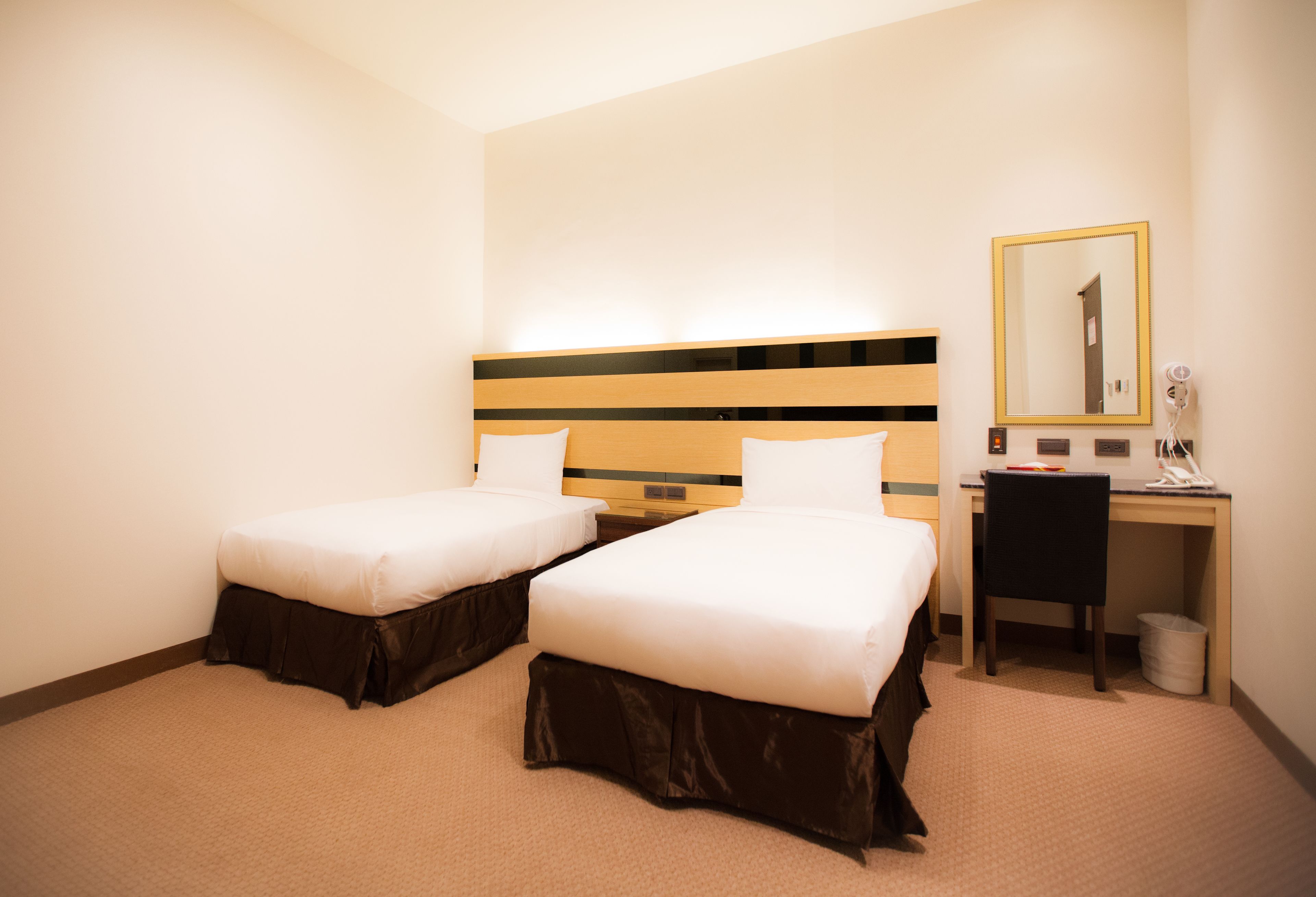 Une chambre d’hôtel avec deux lits simples, un bureau avec une chaise, un miroir et une poubelle.