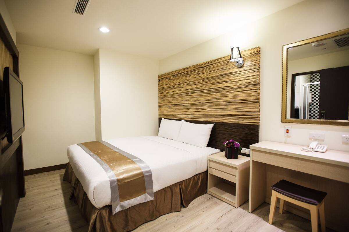 A hotel room with a large bed, a wooden headboard, a bedside table with a phone, a small bench, and a mirror.