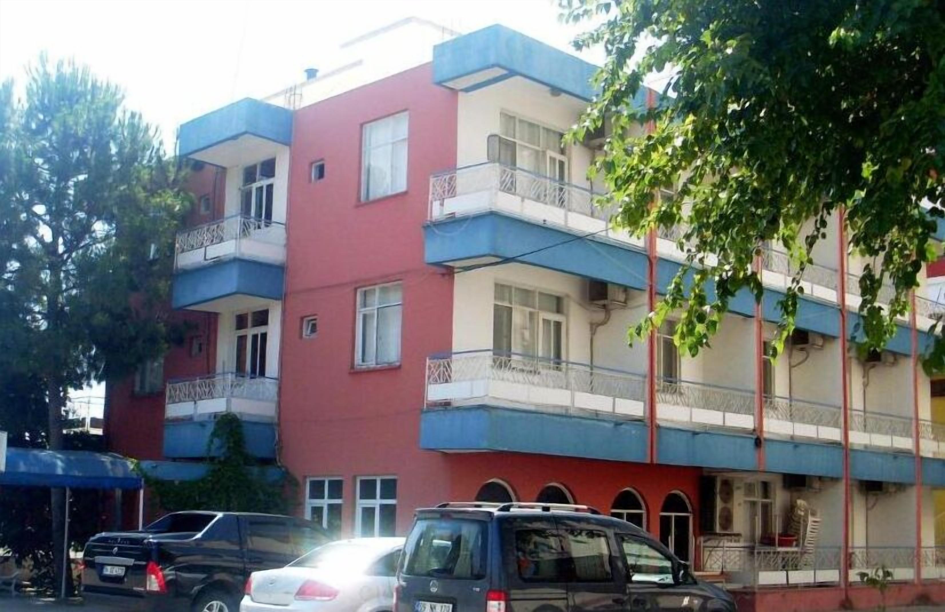 A multi-story residential building with balconies, parked cars, and trees.