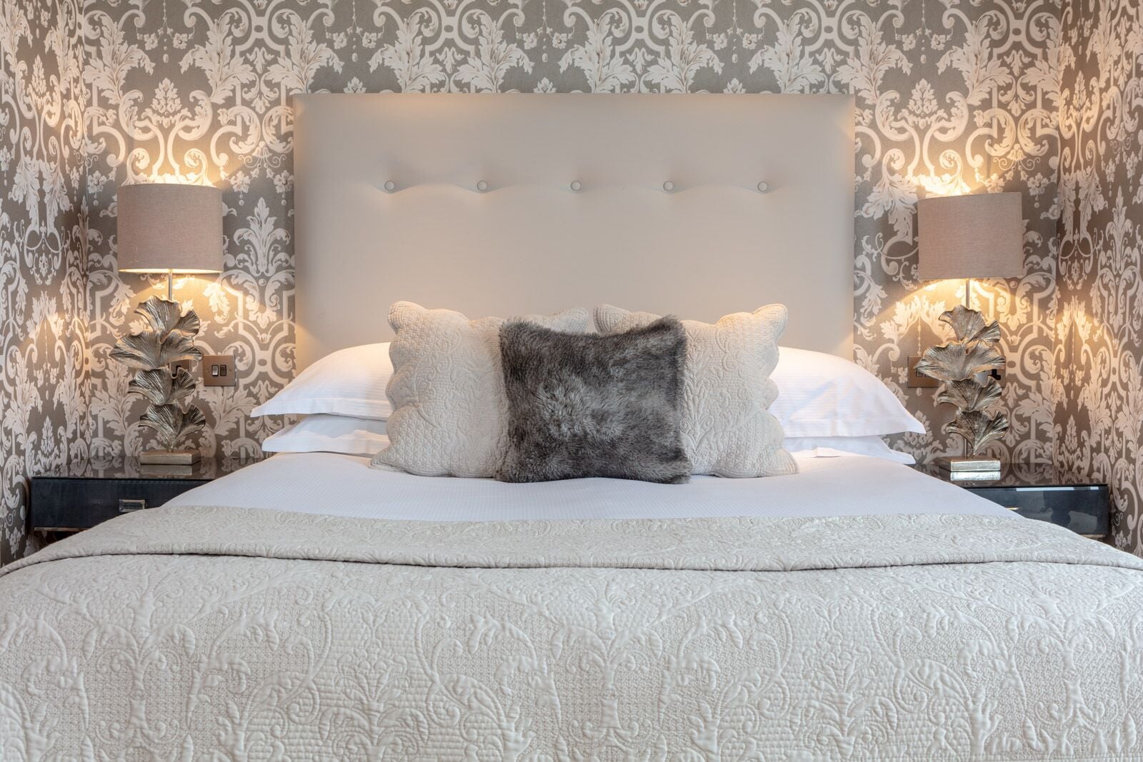 Una cama bien hecha con una colcha estampada y dos almohadas decorativas, flanqueada por lámparas fijadas a la pared y un espejo decorativo.