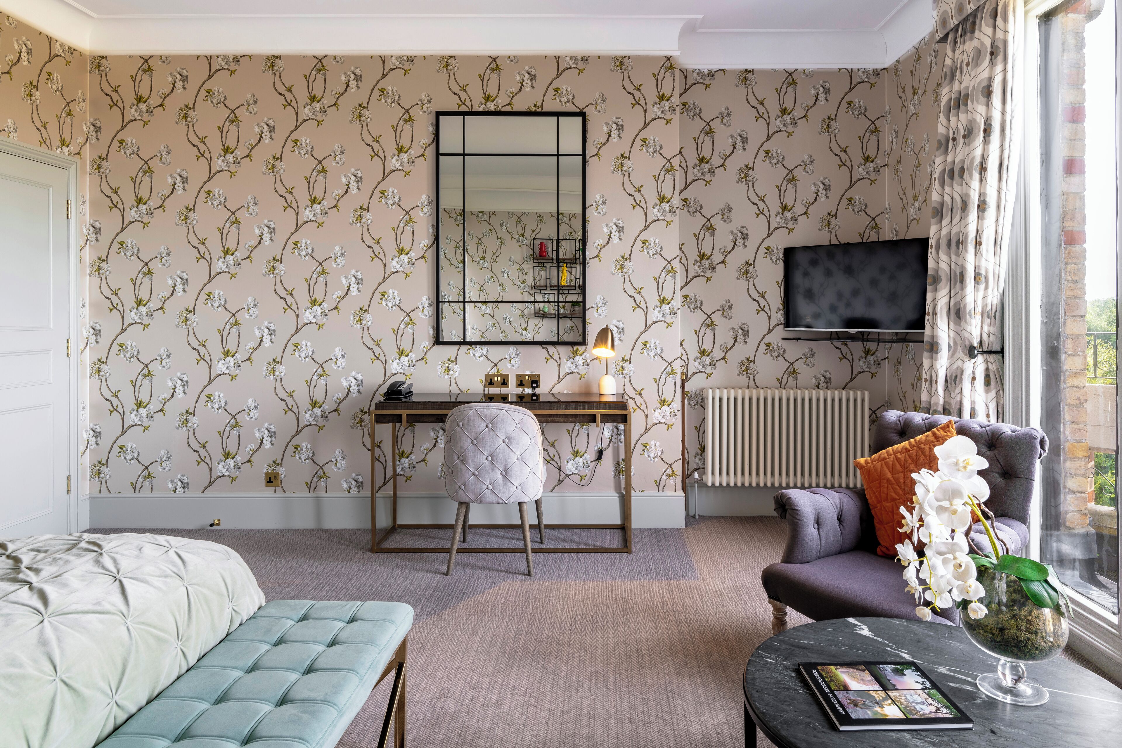 A bedroom with a bed, a desk, a chair, a sofa, a table with a book and a vase, a mirror, and a TV.