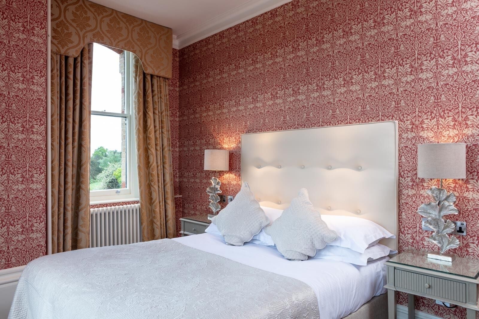 A neatly made bed with a white headboard and two pillows, a window with curtains, and a wall with a floral pattern.