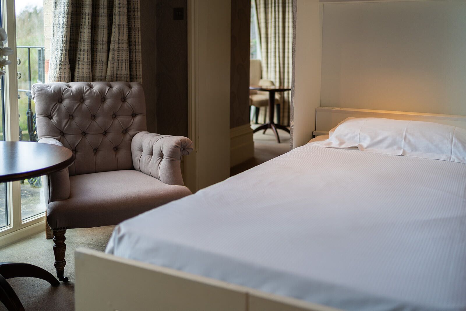 Une chambre d’hôtel avec un lit, un fauteuil capitonné, une petite table ronde et une vue sur l’extérieur par une fenêtre munie de rideaux.