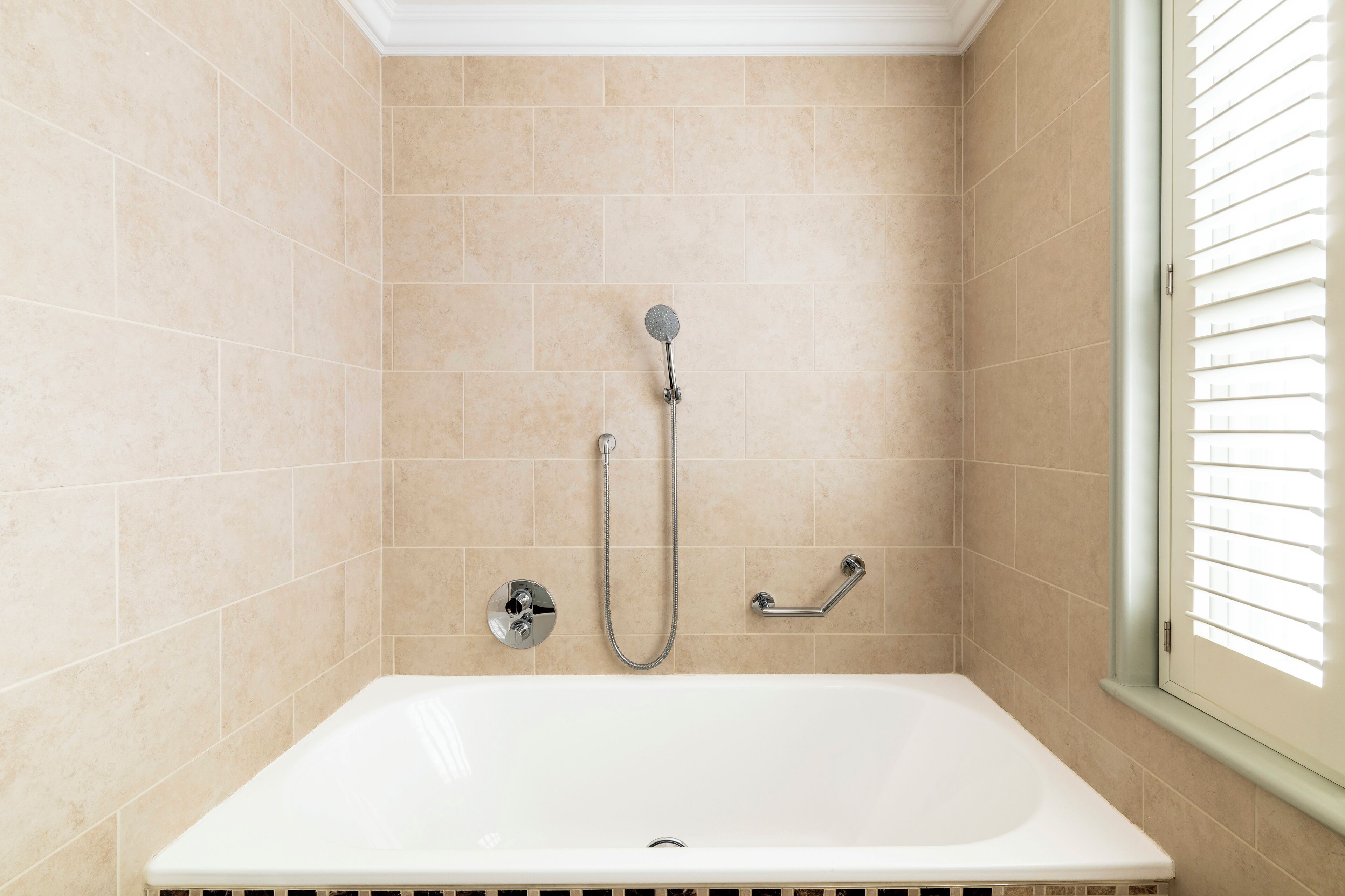 A bathroom with a white bathtub, beige tiles, a showerhead, and a window with blinds.