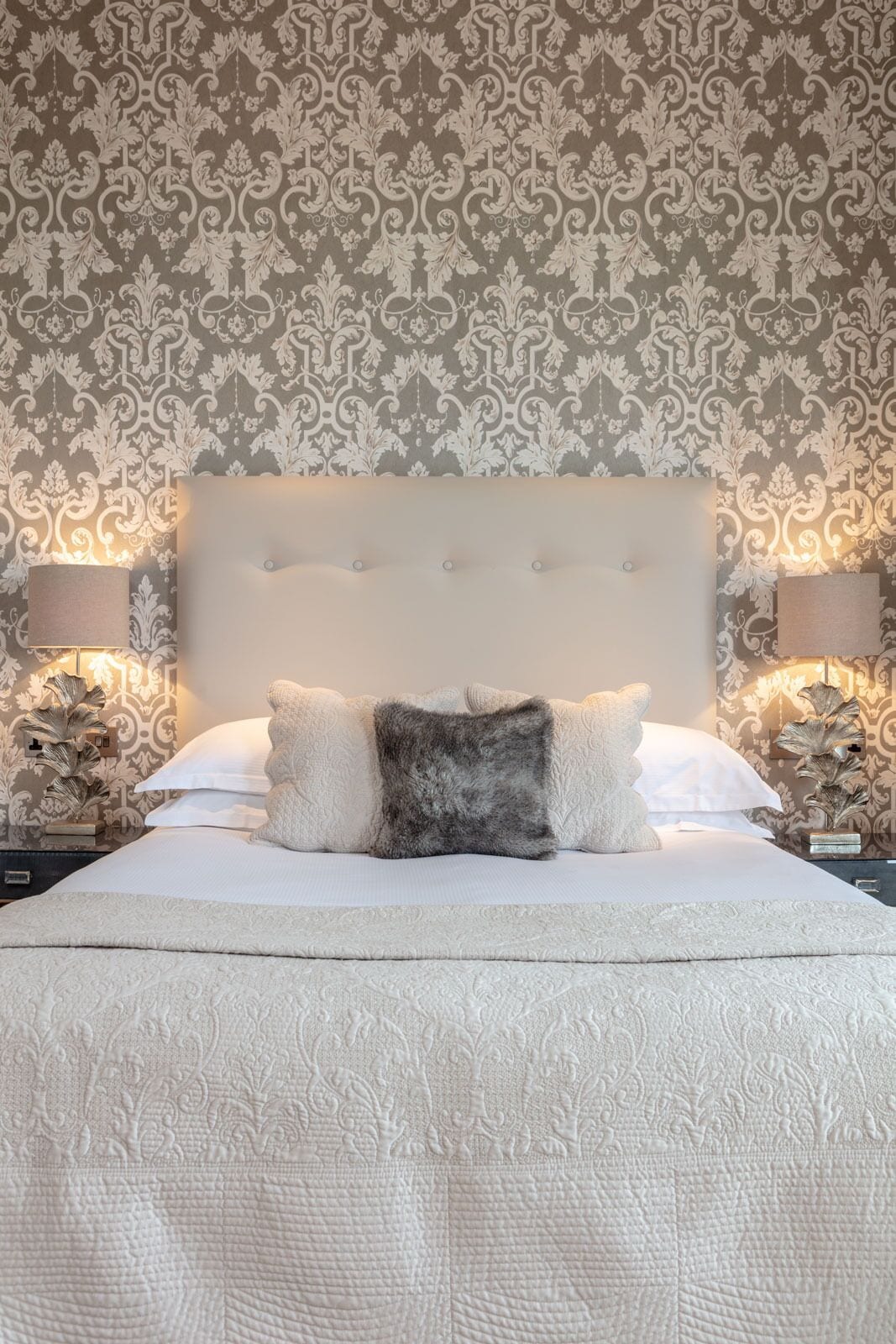 A bed with a tufted headboard, two bedside lamps, and a patterned wallpaper.