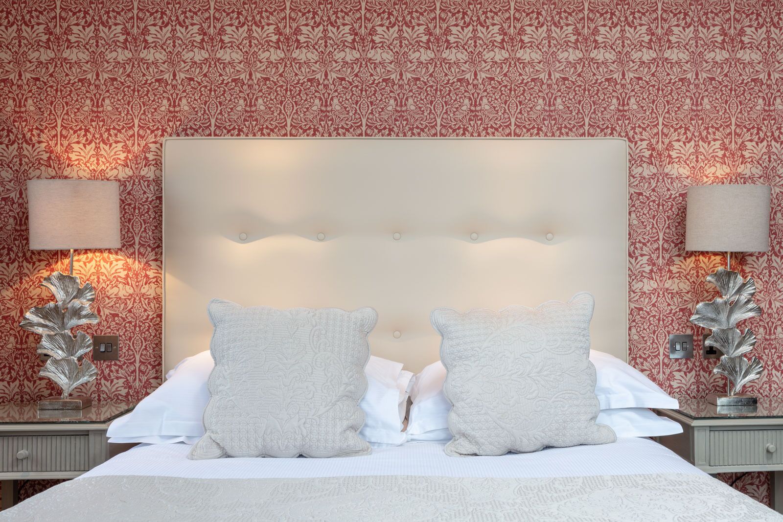 A hotel room with a large bed, two decorative pillows, a tufted headboard, and patterned wallpaper.