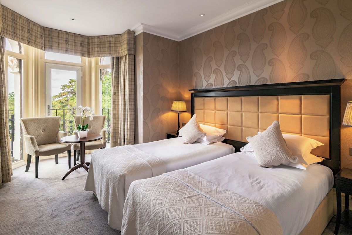 A hotel room with two beds, a small round table, a chair, and a view of the outdoors.
