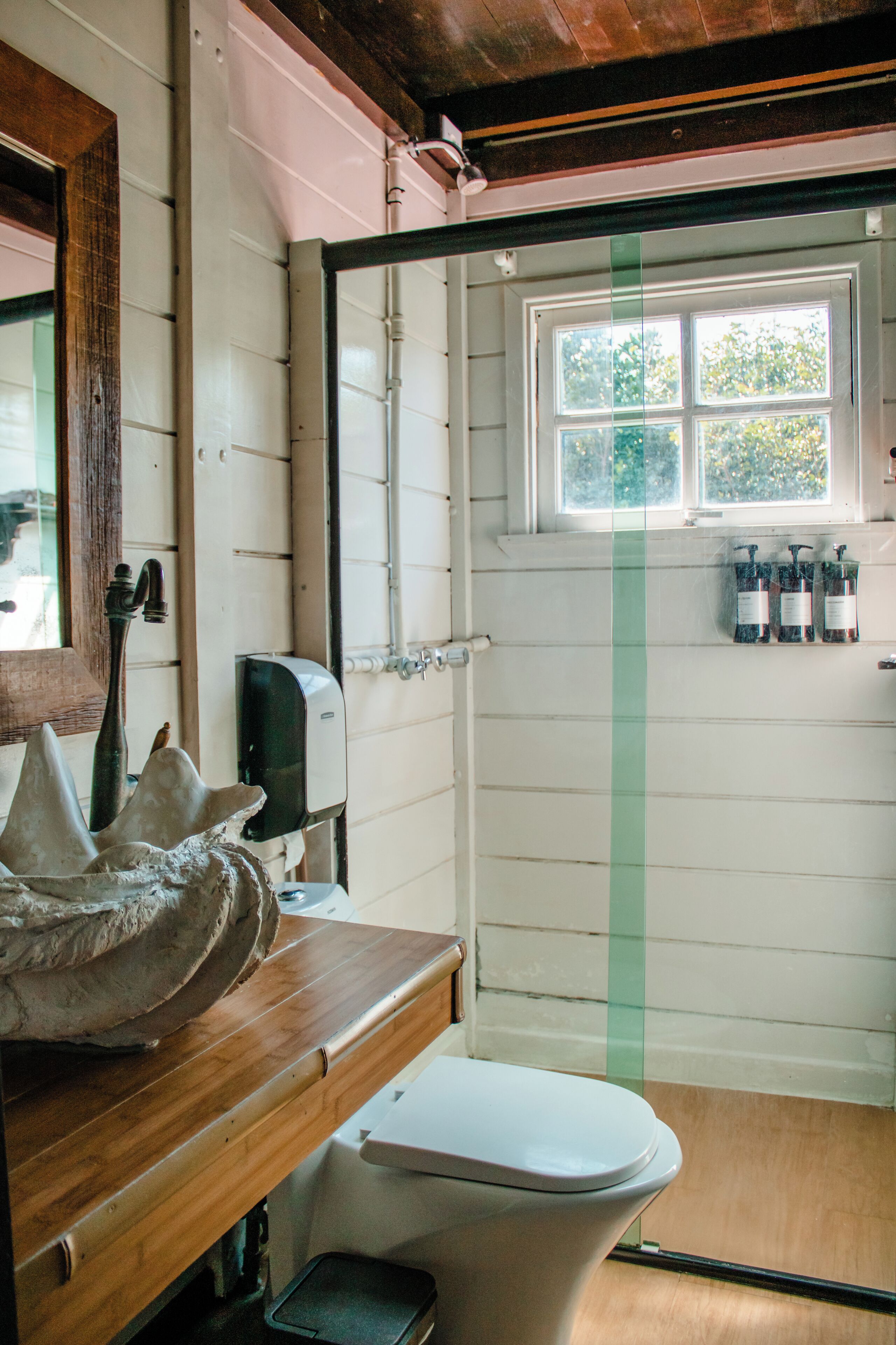 A bathroom with a wooden vanity, a toilet, a window with a view of greenery, and a hand soap dispenser on the wall.