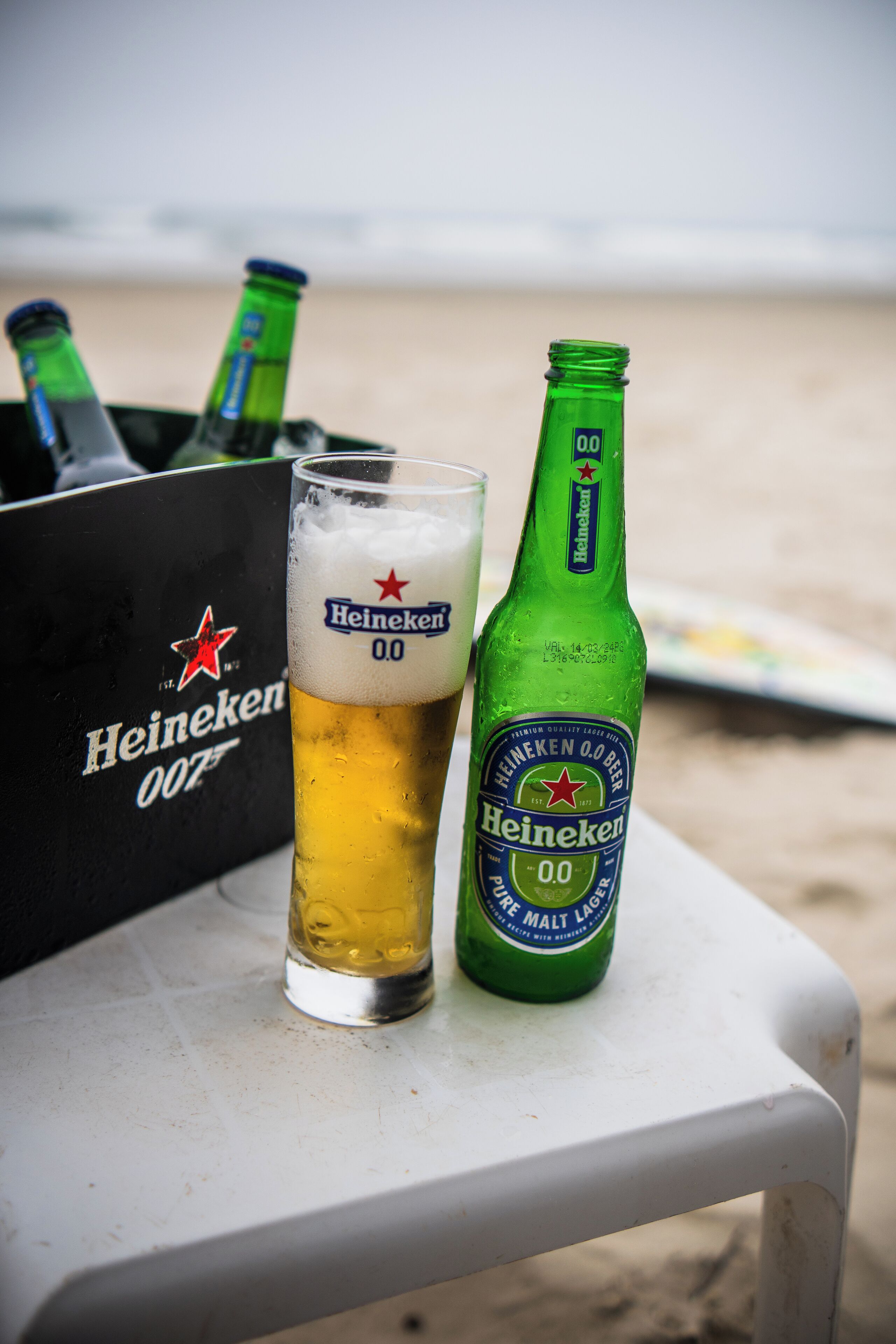 A Heineken beer bottle and glass on a white plastic chair at the beach.