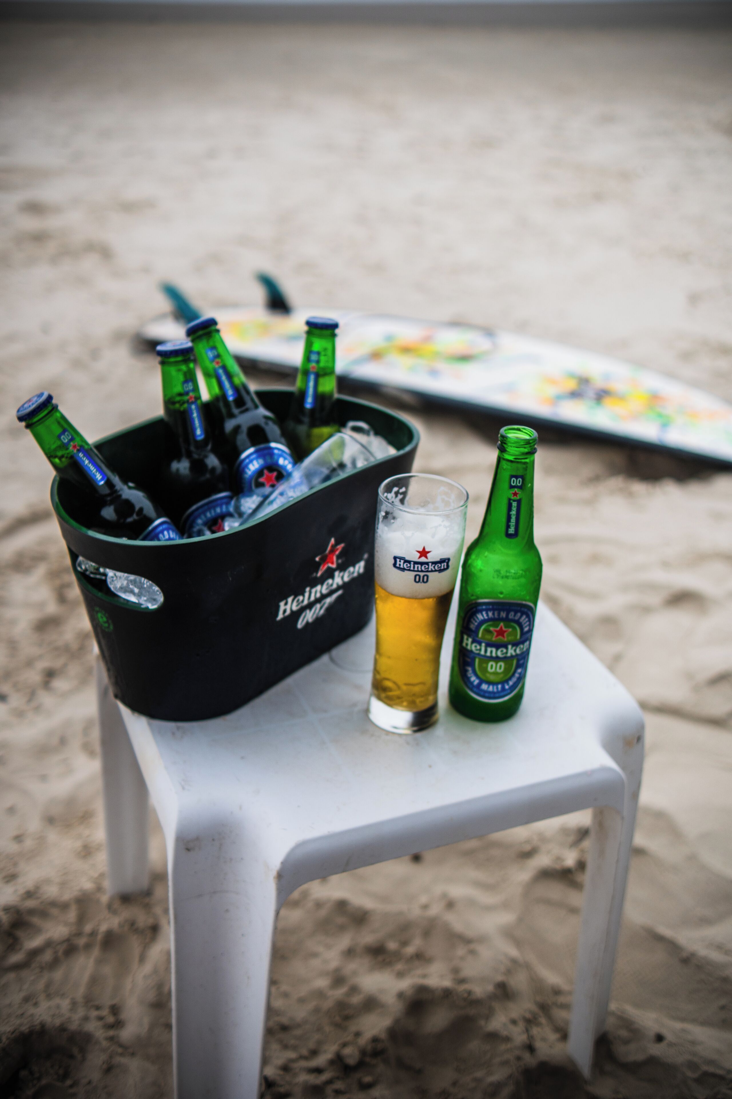 A beach scene with a black Heineken cooler filled with bottles and a glass on a white plastic chair.