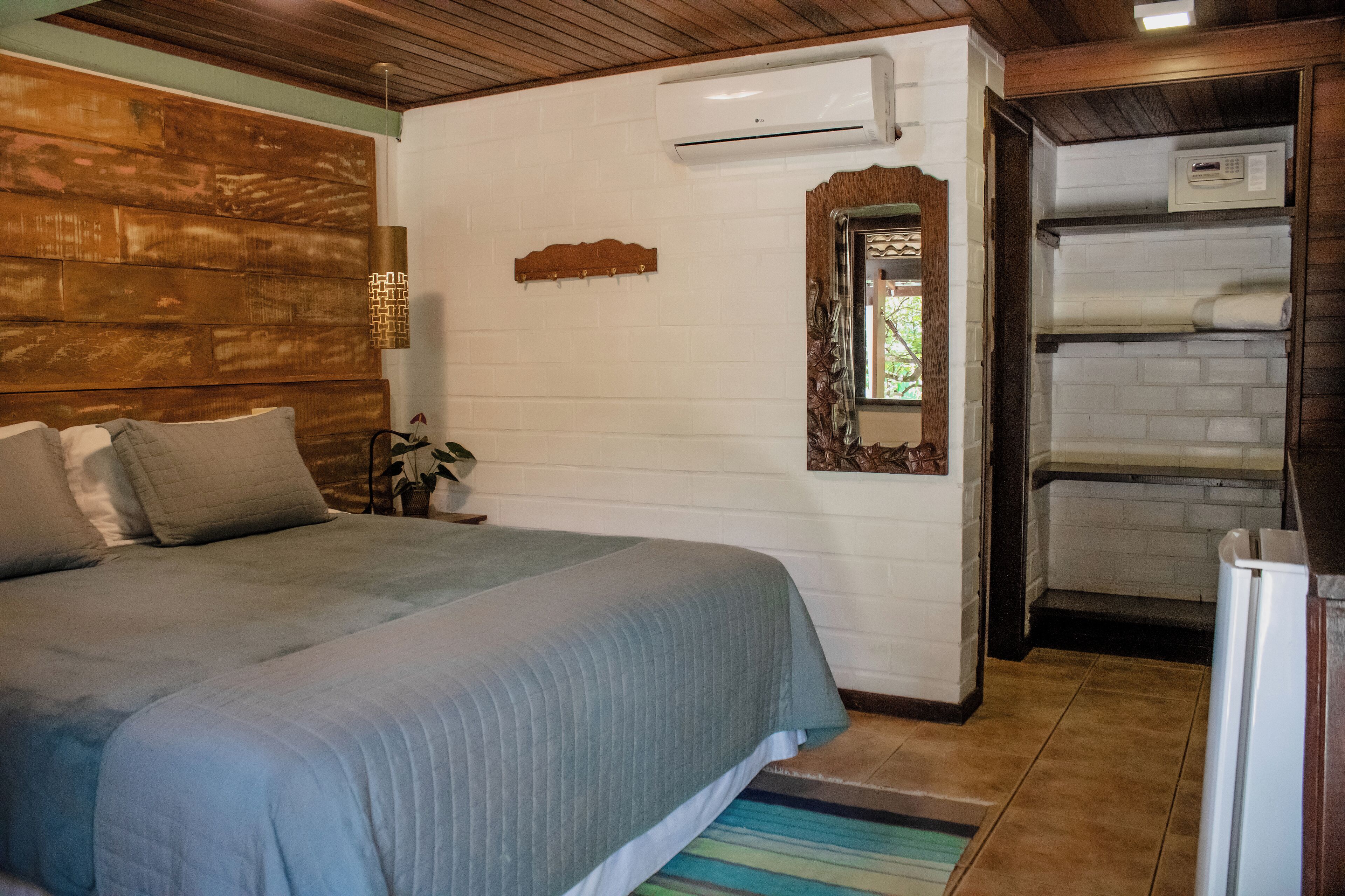 Un dormitorio con cama, cabecero de madera, aire acondicionado de pared, espejo y una pequeña estantería con una planta.