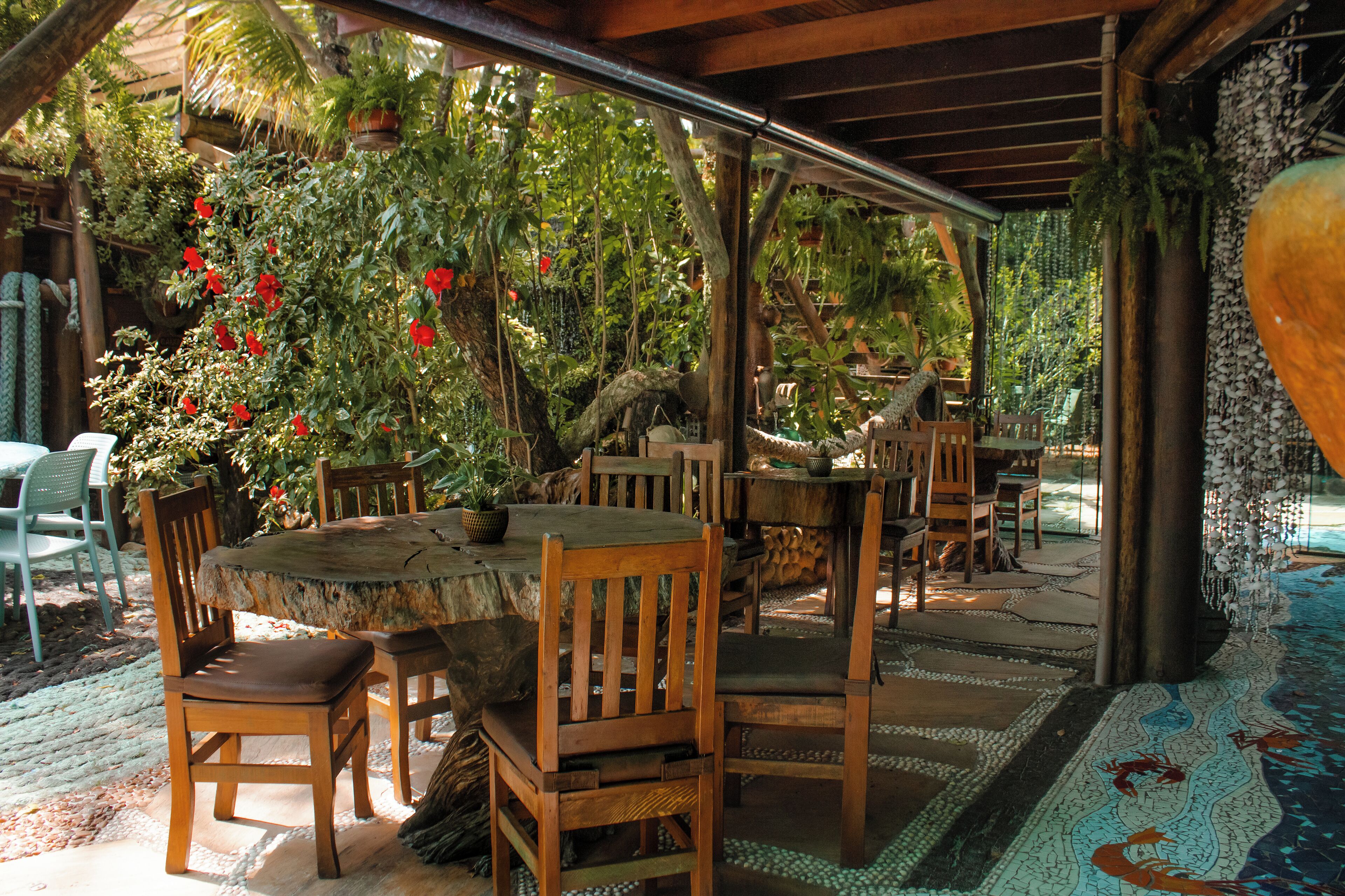 An outdoor patio with wooden tables and chairs, surrounded by lush greenery and red flowers.