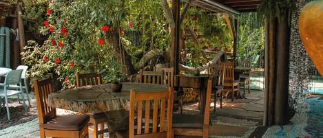 An outdoor patio with wooden tables and chairs, surrounded by lush greenery and red flowers.