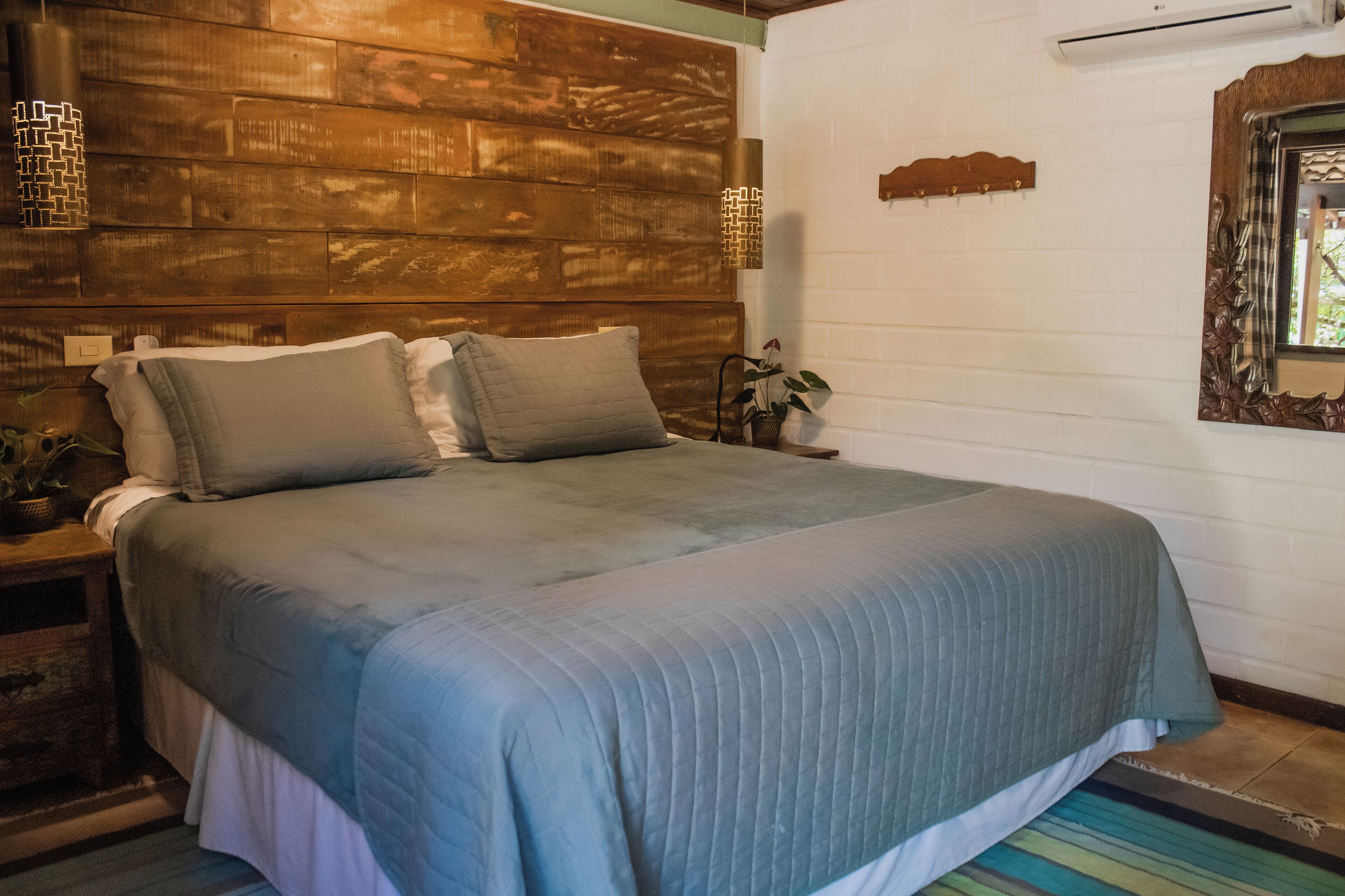 A bedroom with a wooden headboard, a bed with grey bedding, a bedside table with a lamp, and a potted plant.