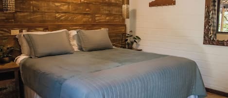 A bedroom with a wooden headboard, a bed with grey bedding, a bedside table with a lamp, and a potted plant.